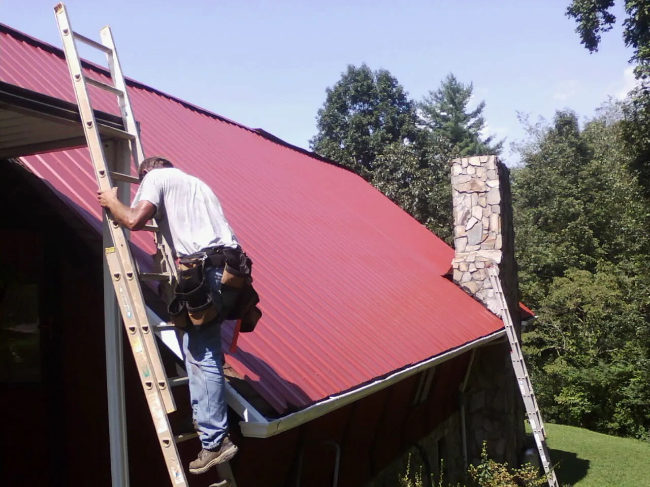Roofer on a ladder beside a red metal roof and stone chimney — Soffit & Fascia Repair by Roofing Brothers in Trinity